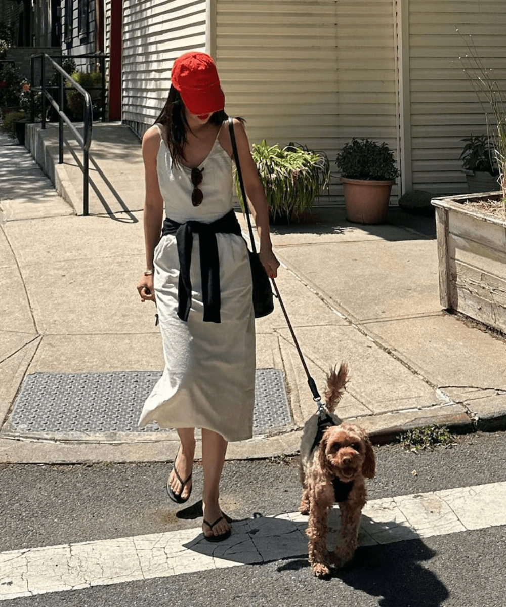 Mimi Nguyen - vestido midi branco, boné vermelho e tricot amarrado na cintura - roupas brancas - primavera - mulher de boné andando na rua com um cachorro ao lado - https://stealthelook.com.br