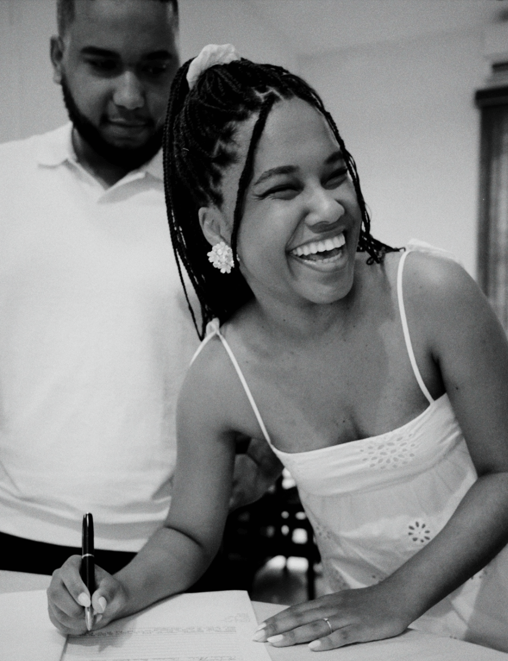 Clau Ribeiro e Gabriel Menezes - vestido curto branco - fotos no cartório - verão - foto em preto e branco mostra mulher de tranças sorrindo assinando um papel - https://stealthelook.com.br