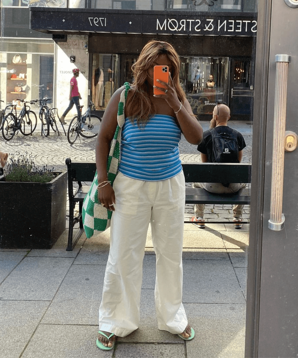 Nnenna Echem - calça pantalona branca, chinelo e top listrado azul sem alças - looks fresquinhos - verão - mulher negra em pé na rua tirando foto em frente a um espelho - https://stealthelook.com.br