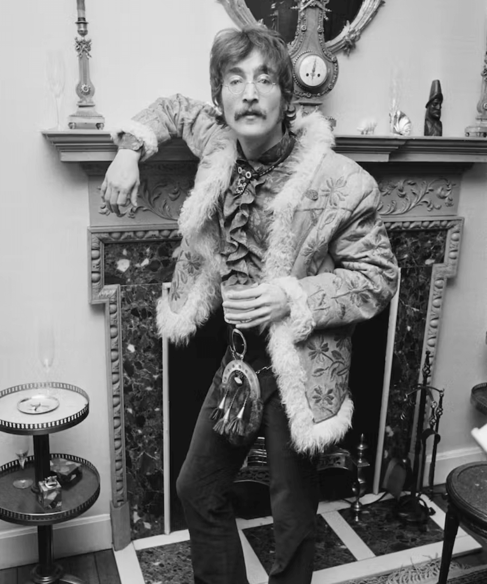 John Lennon - Casaco penny lane, calças e botas - tendência do inverno 2024 - inverno - homem de bigode parado em pé em frente a uma lareira - https://stealthelook.com.br