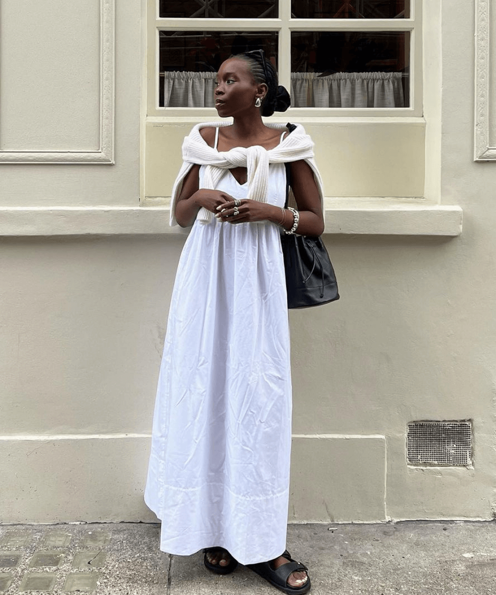 Danielle Oreoluwa Jinadu - vestido branco longo, papete preta, acessórios e tricot amarrado nos ombros - looks estilosos - verão - mulher negra em pé na rua olhando para o lado - https://stealthelook.com.br