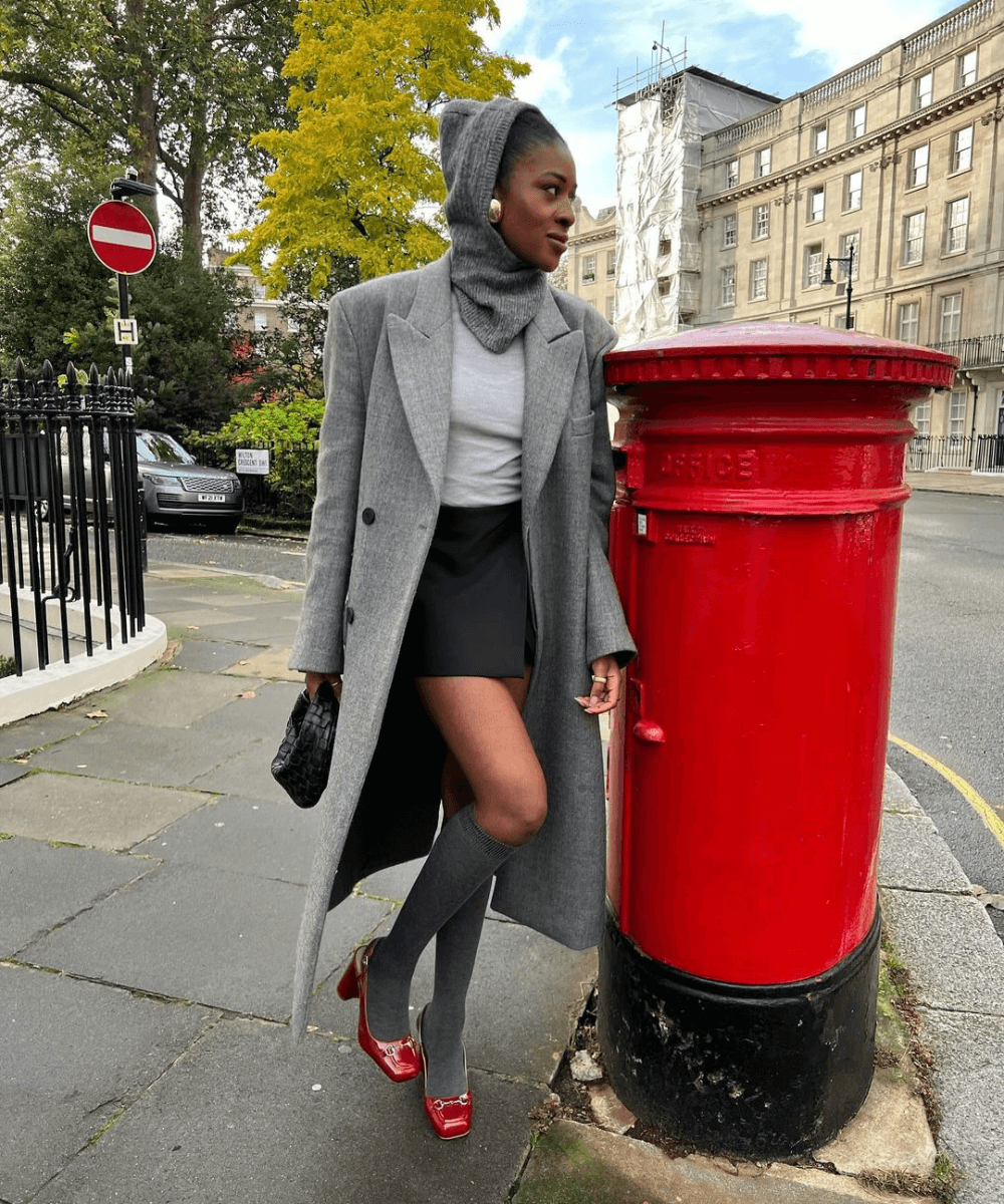 @georgia_3.0 - minissaia, meias cinzas, slingback vermelho, casaco longo cinza - slingbacks - inverno - mulher negra em pé na rua com a perna dobrada - https://stealthelook.com.br