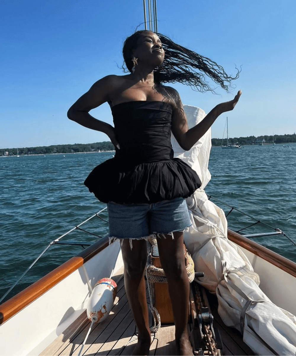Tahirah Hairston - shorts jeans e blusa preta em peplum - tendências de moda - verão - mulher negra em pé rindo em um barco no mar - https://stealthelook.com.br