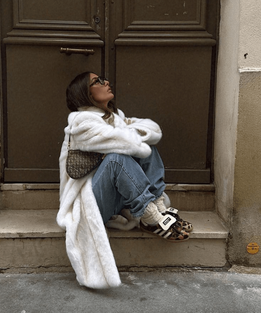 @cestclau - calça jeans, tênis oncinha adidas, t-shirt branca e casaco longo peludo - tendência do inverno - inverno - mulher sentada de lado com as pernas dobradas na rua - https://stealthelook.com.br