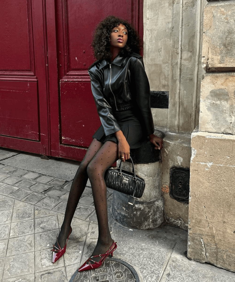 Emmanuelle Koffi - minissaia preta, meia-calça, slingback vermelho e jaqueta de couro preta - sapato vermelho - inverno - mulher negra sentada em um banco na rua olhando para o lado - https://stealthelook.com.br