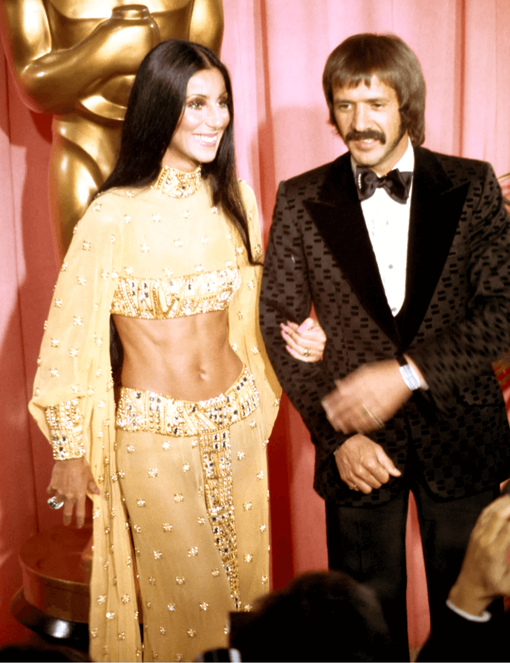 Cher e Bono - top cropped de manga longa e saia longa em tons de dourado - Looks da Cher - verão - mulher e homem juntos parados sorrindo - https://stealthelook.com.br