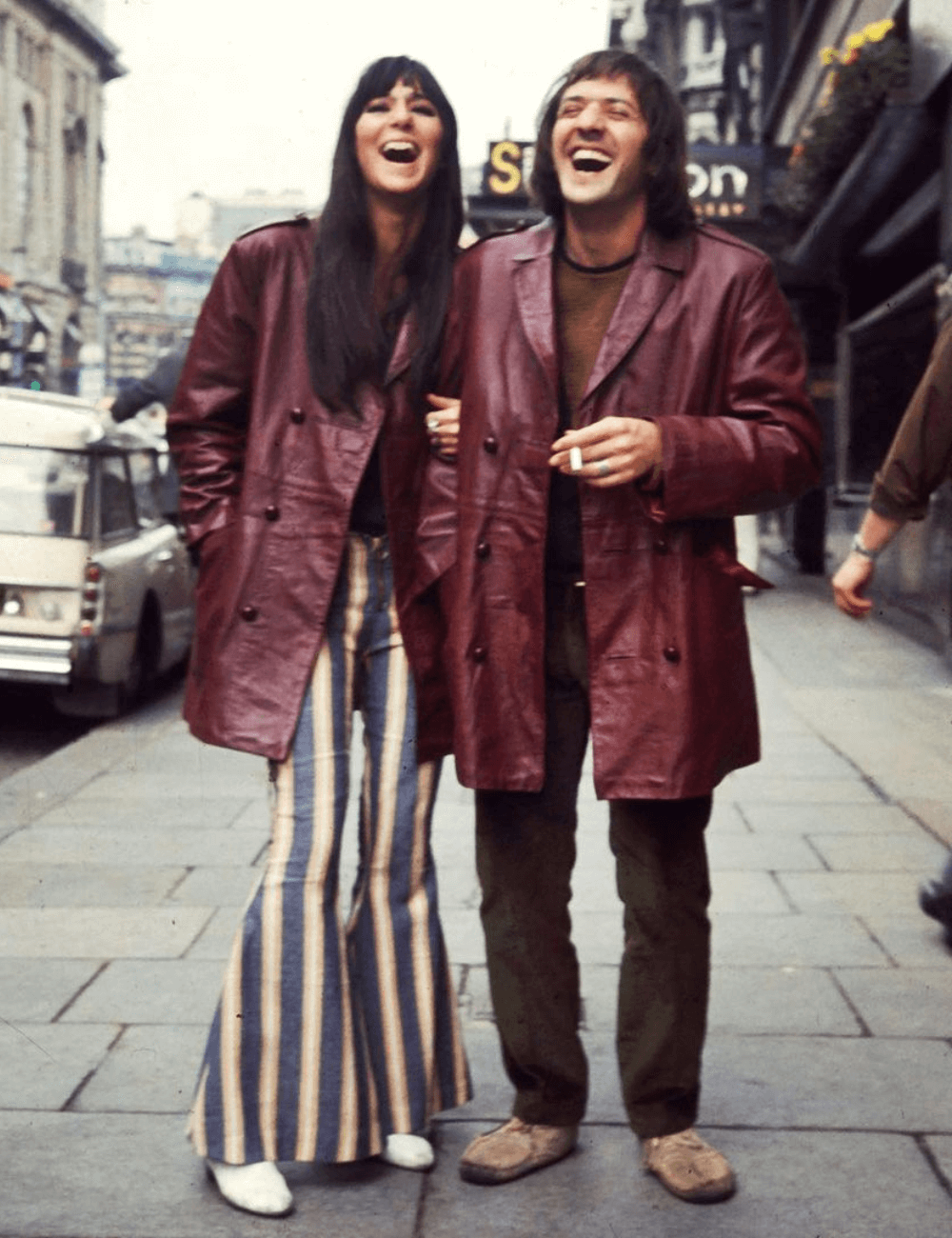 Cher e Sonny - calça boca de lino listrada, jaqueta de couro vermelha escuro  e botas - Looks da Cher - inverno - mulher em pé sorrindo na rua de braços dados com o parceiro - https://stealthelook.com.br