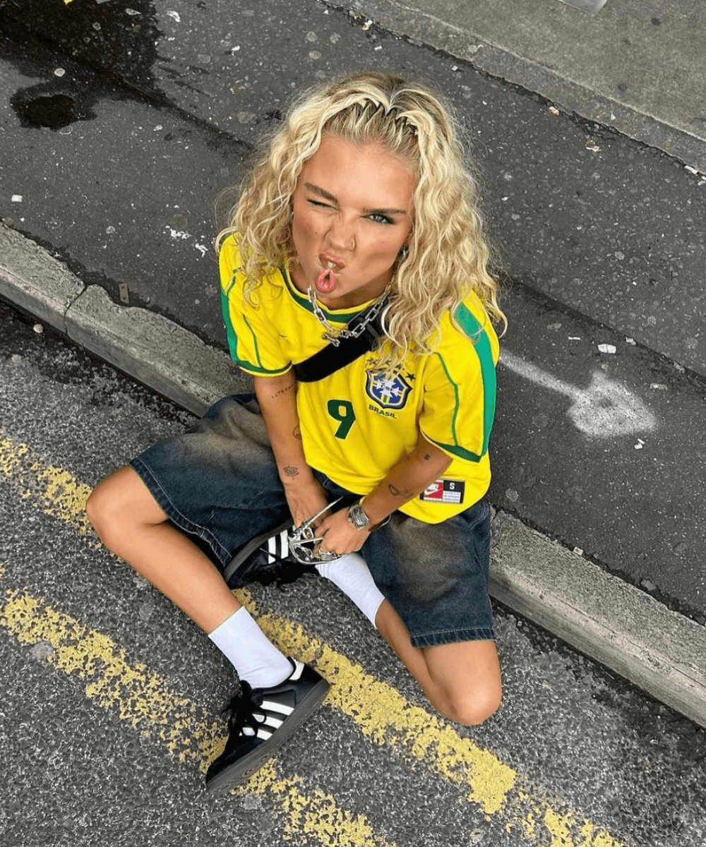 Daisy Birchall - camisa da seleção brasileira, bermuda e adidas samba - moda e futebol - verão - mulher loira sentada no chão da rua olhando para câmera - https://stealthelook.com.br