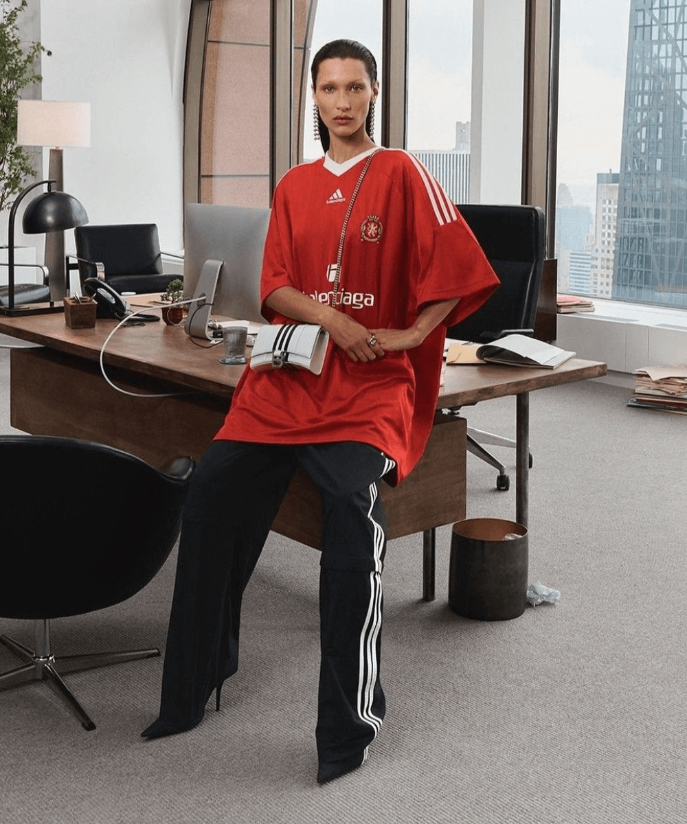 Bella Hadid - blusa de futebol vermelha, calça preta e salto alto - moda e futebol - inverno - mulher sentada em uma mesa - https://stealthelook.com.br