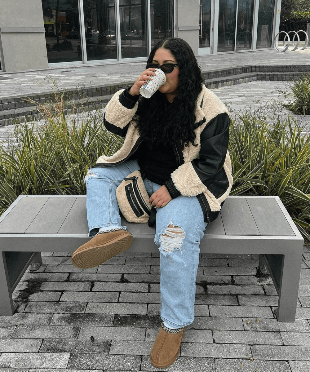@stephmnavarro - calça jeans, ugg, blusa preta e jaqueta shearling preta - sapatos de inverno - inverno - mulher de óculos sentada em um banco na rua com uma perna dobrada - https://stealthelook.com.br
