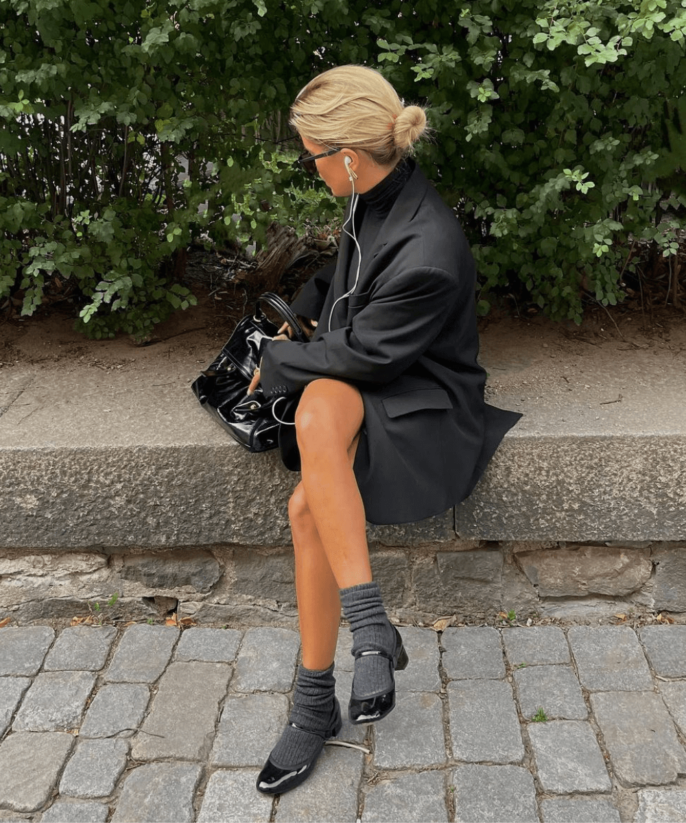 Matilda Djerf - blazer oversized, meias cinza, sapato boneca preto de salto - sapatos de inverno - inverno - mulher loira de fones sentada na rua  - https://stealthelook.com.br