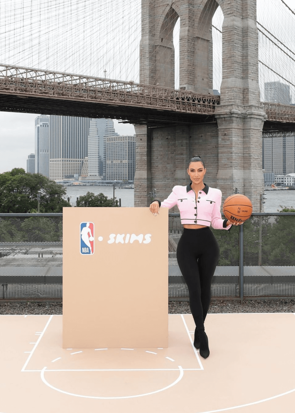 Kim Kardashian - Calça preta, salto alto e casaco rosa - moda e basquete - inverno - mulher em pé segurando uma bola de basquete na mão - https://stealthelook.com.br