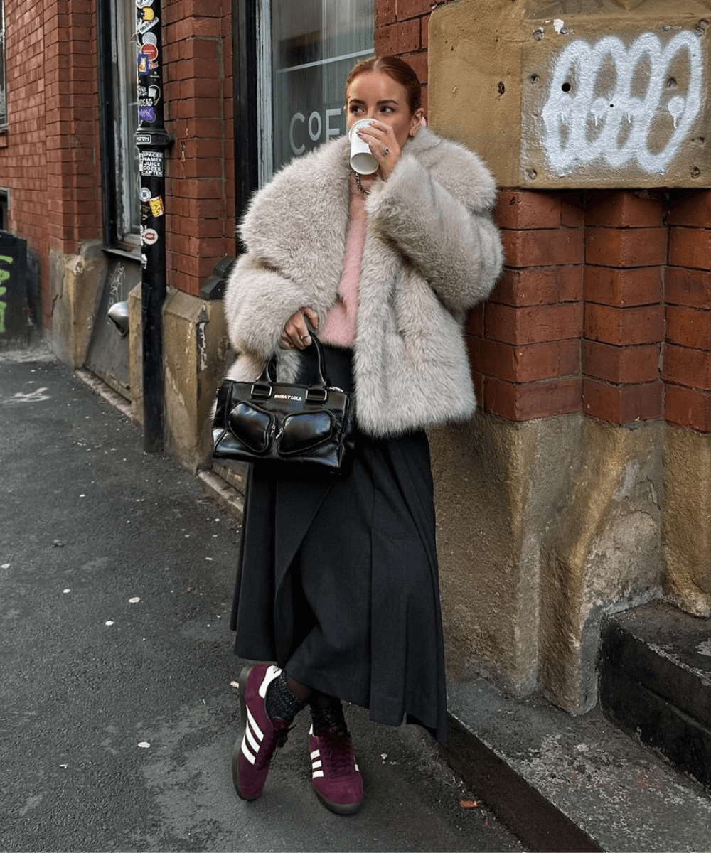 @rebeccaferrazwyatt - casaco de pele fake rosa, saia preta, tênis adidas e bolsa vermelha - tendências do inverno 2024 - inverno - mulher loira em pé na rua bebendo café - https://stealthelook.com.br