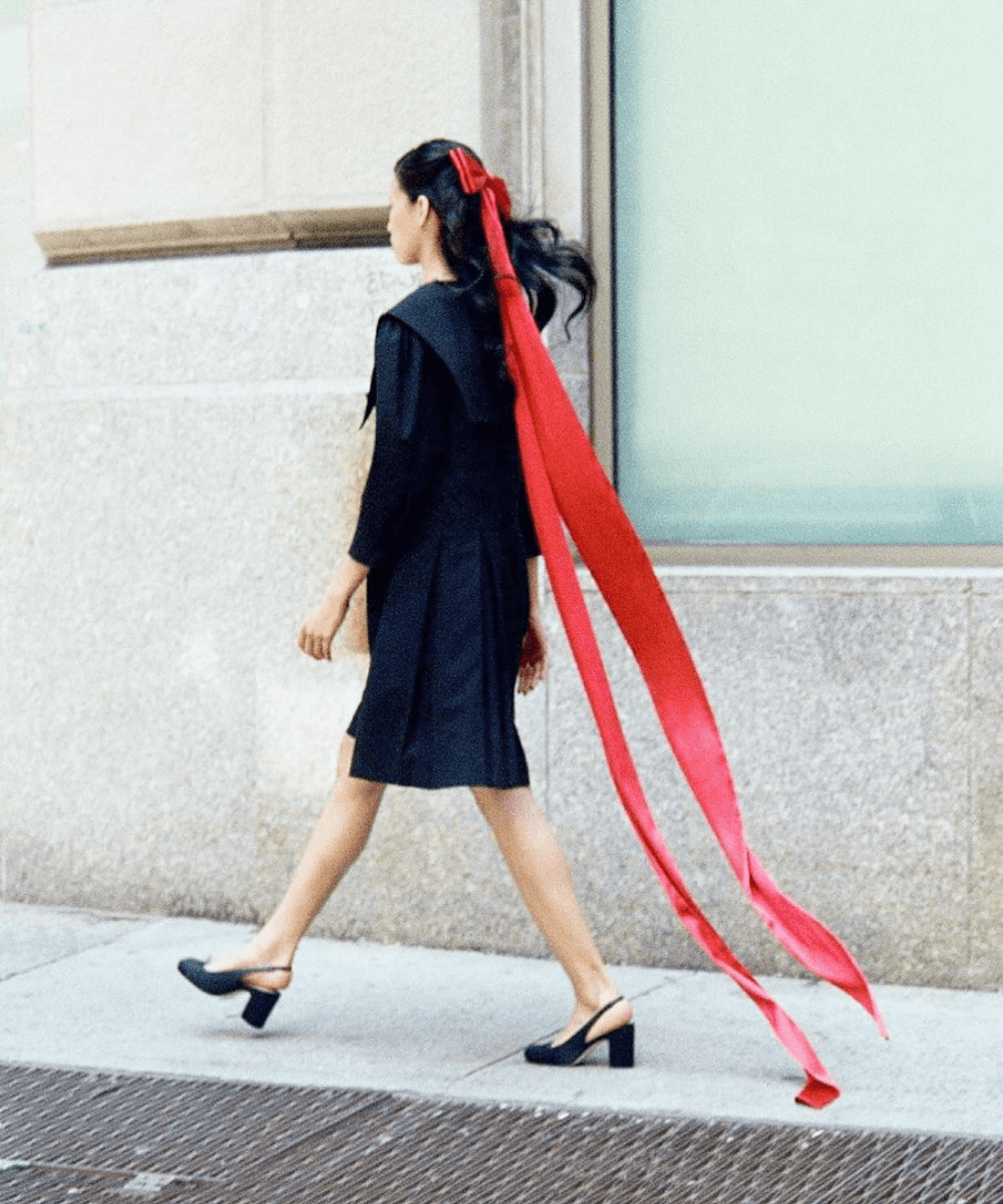 Sandy Liang - vestido preto, sapato boneca e laço vermelho - Sandy Liang - inverno - mulher andando na rua usando um laço gigante vermelho preso no cabelo - https://stealthelook.com.br