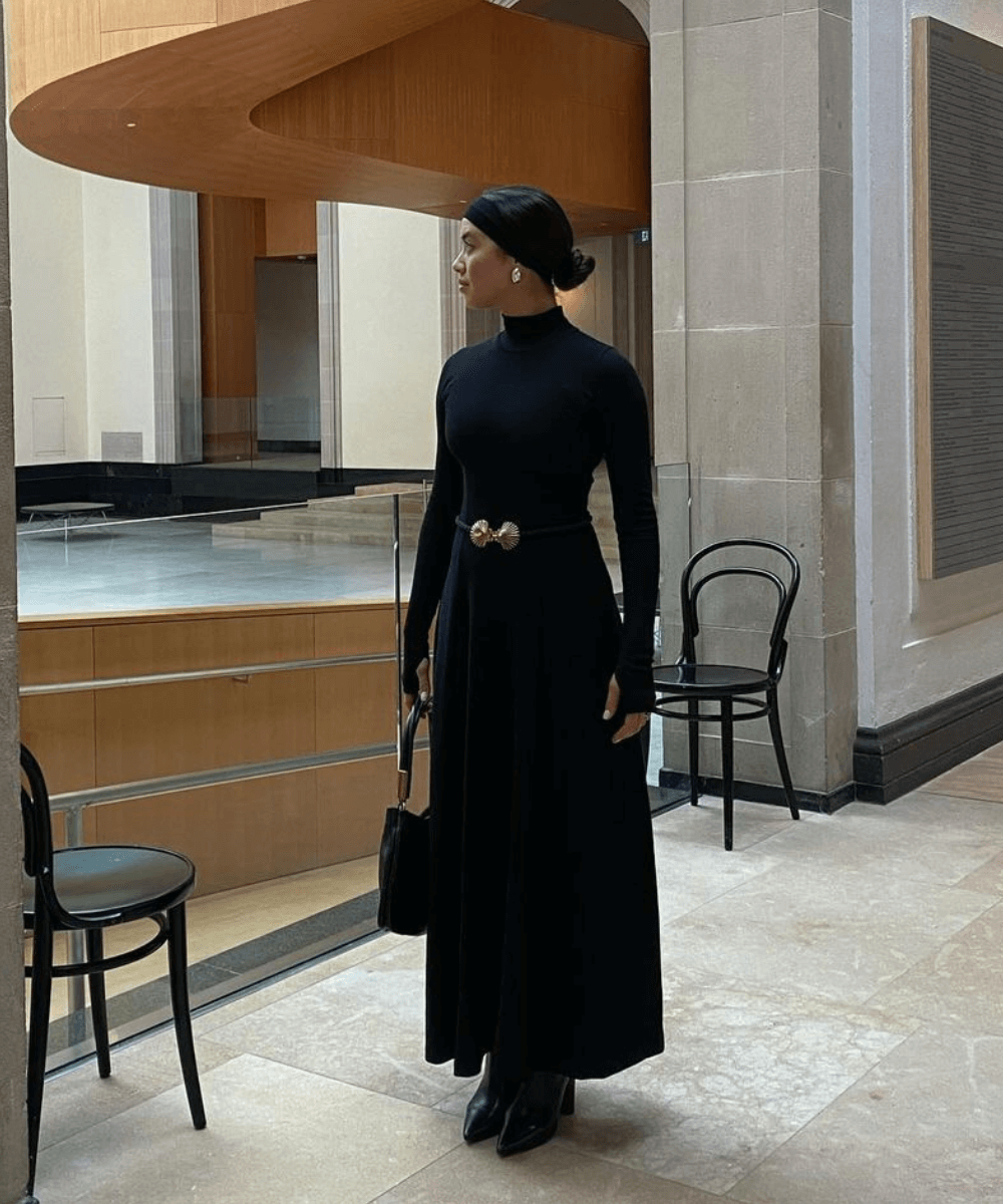 Sasha Mei - saia longa preta, blusa preta e botas - acessório tendência - inverno - mulher em pé em uma sala - https://stealthelook.com.br