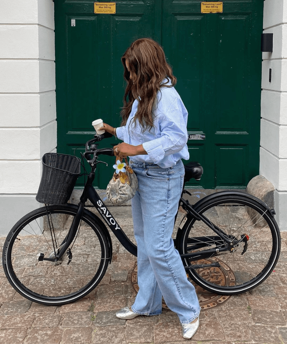 Nnenna Echem - calça jeans, sapatilha prata e camisa azul - armário cápsula - verão - mulher negra em pé na rua ao lado de uma bicicleta - https://stealthelook.com.br