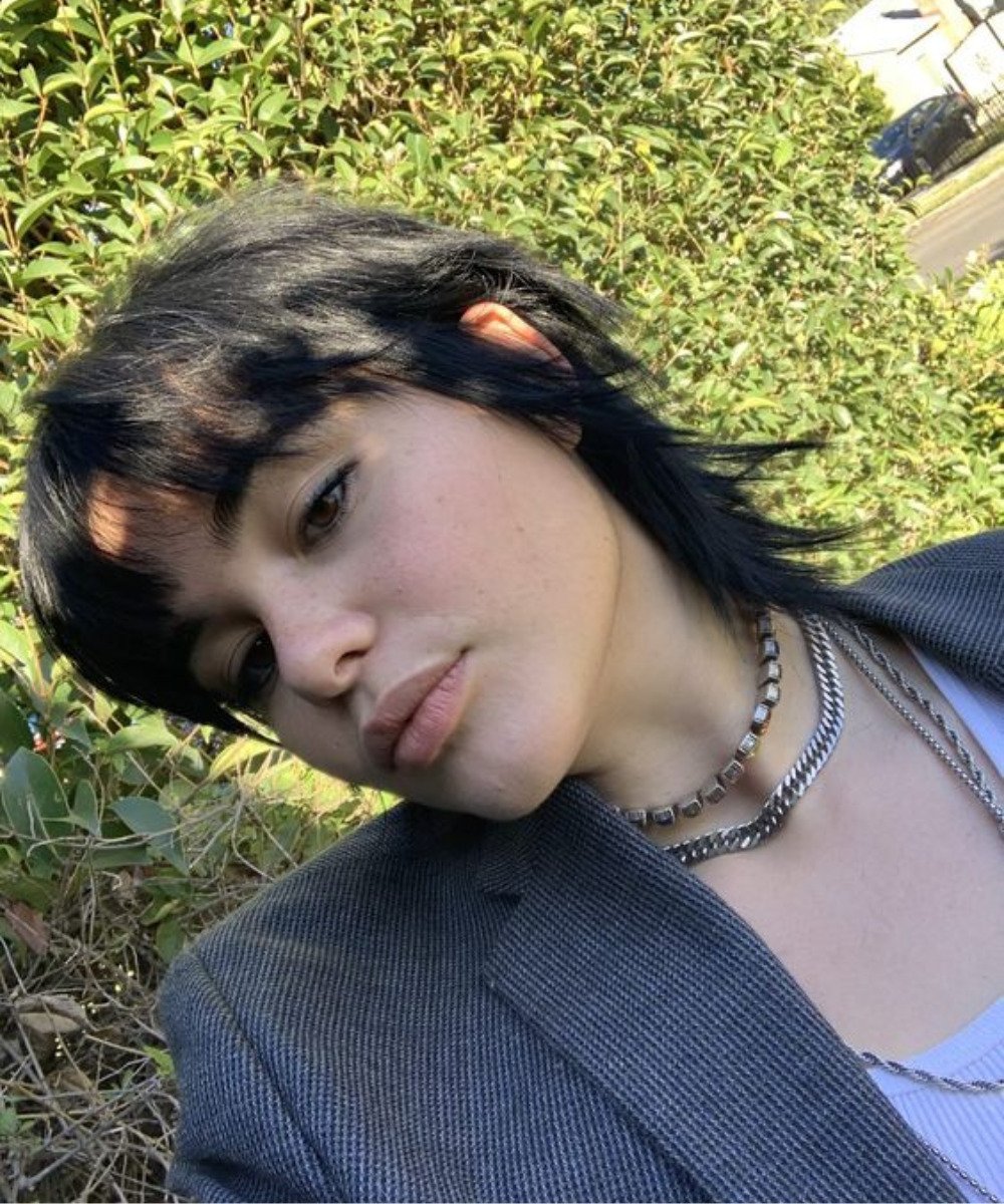 Mulher com cabelo estilo mullet feminino, usando terno e colares, em um ambiente ao ar livre.