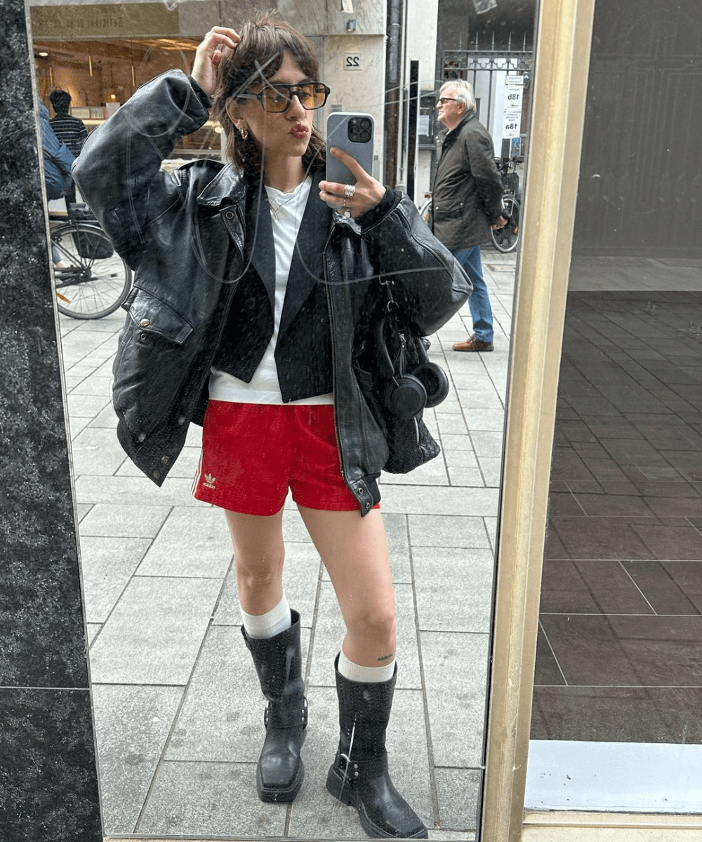 Rebeca Oksana - short vermelho, blusa branca, biker boots e jaqueta de couro - biker boots - outono - mulher na rua usando óculos e tirando foto em frente a uma vitrine - https://stealthelook.com.br