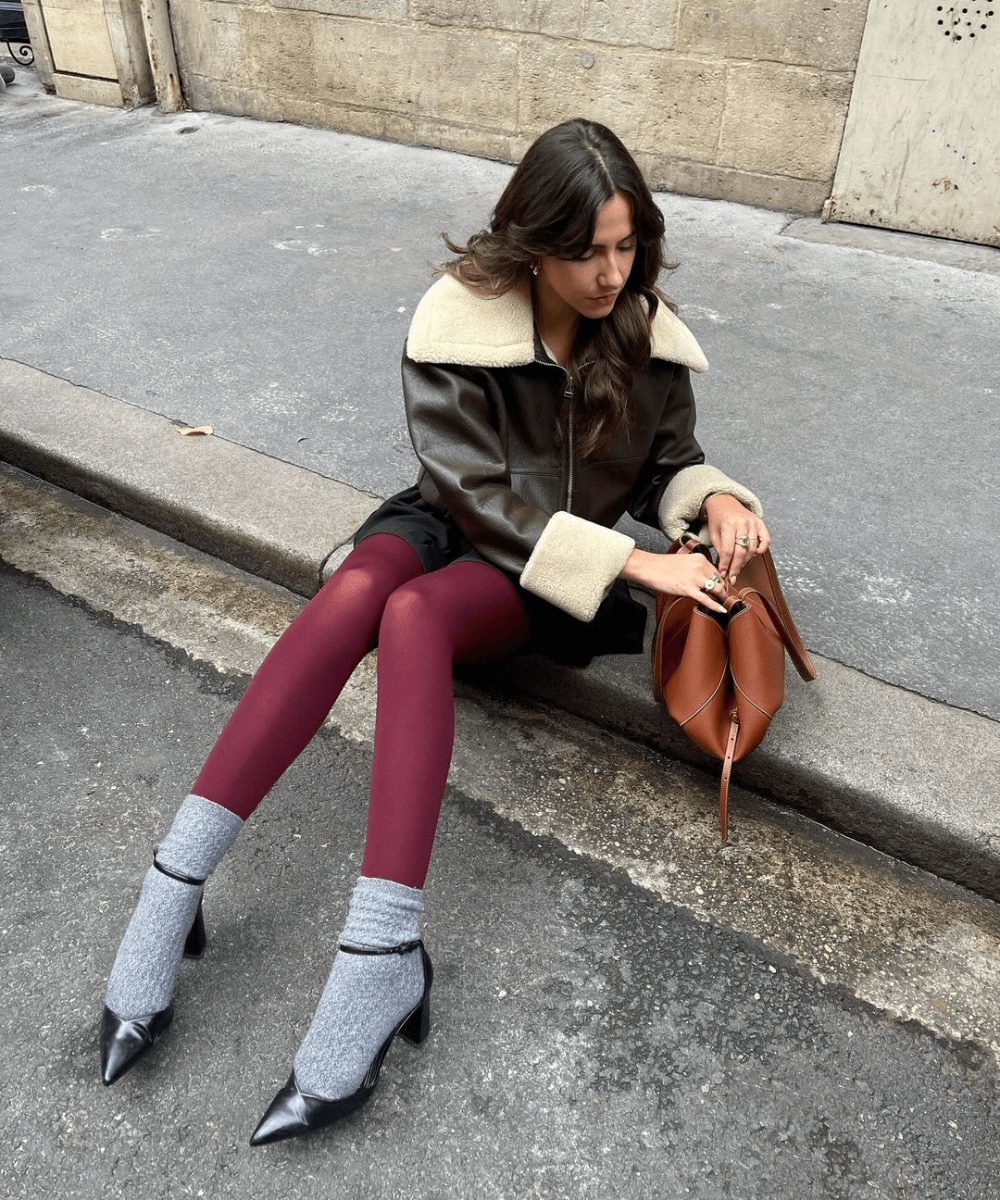 @florettenacer - meia-calça vermelha, meia curta cinza, sapato preto e jaqueta de couro - inverno 2024 - inverno - mulher sentada na rua mexendo na bolsa - https://stealthelook.com.br