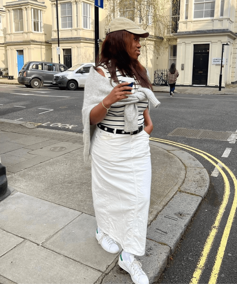 Nnenna Echem - saia midi branca jeans, blusa listrada, boné e tênis branco - looks com tênis - outono - mulher negra andando na rua usando óculos de sol e boné - https://stealthelook.com.br