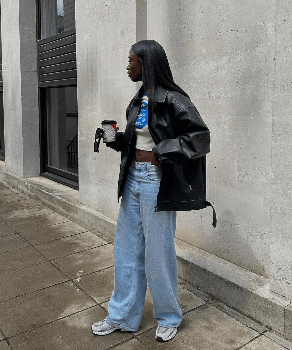 Liv Madeline - calça jeans, regata cropped, jaqueta bomber de couro e tênis esportivos - looks fáceis - outono - mulher negra em pé na rua segurando um café - https://stealthelook.com.br