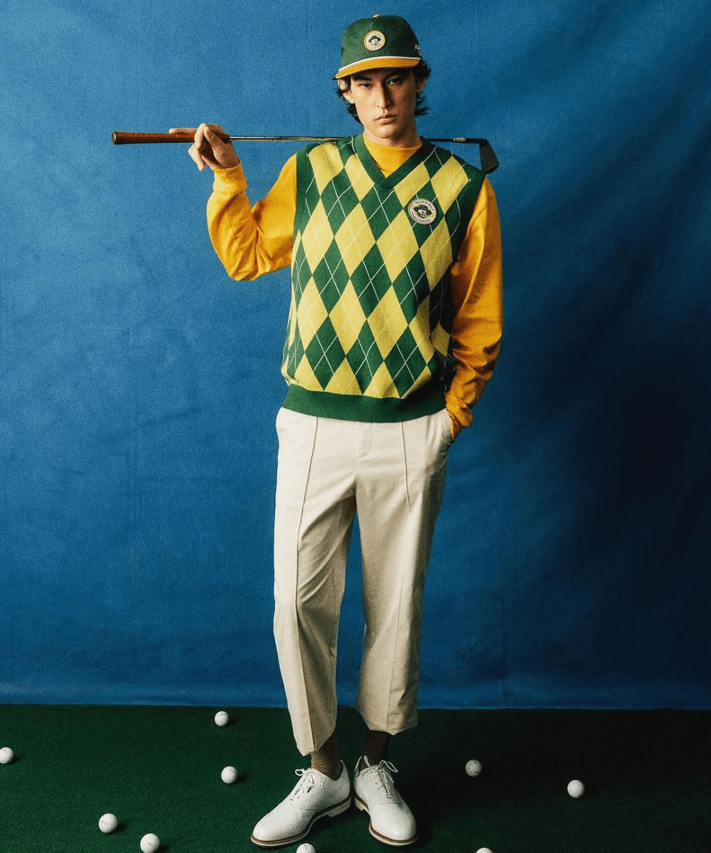 @malbongolf - calça cáqui, colete xadrez e camisa amarela social - moda e golfe - inverno - homem posando em frente a um fundo azul com um taco de golfe nas mãos - https://stealthelook.com.br