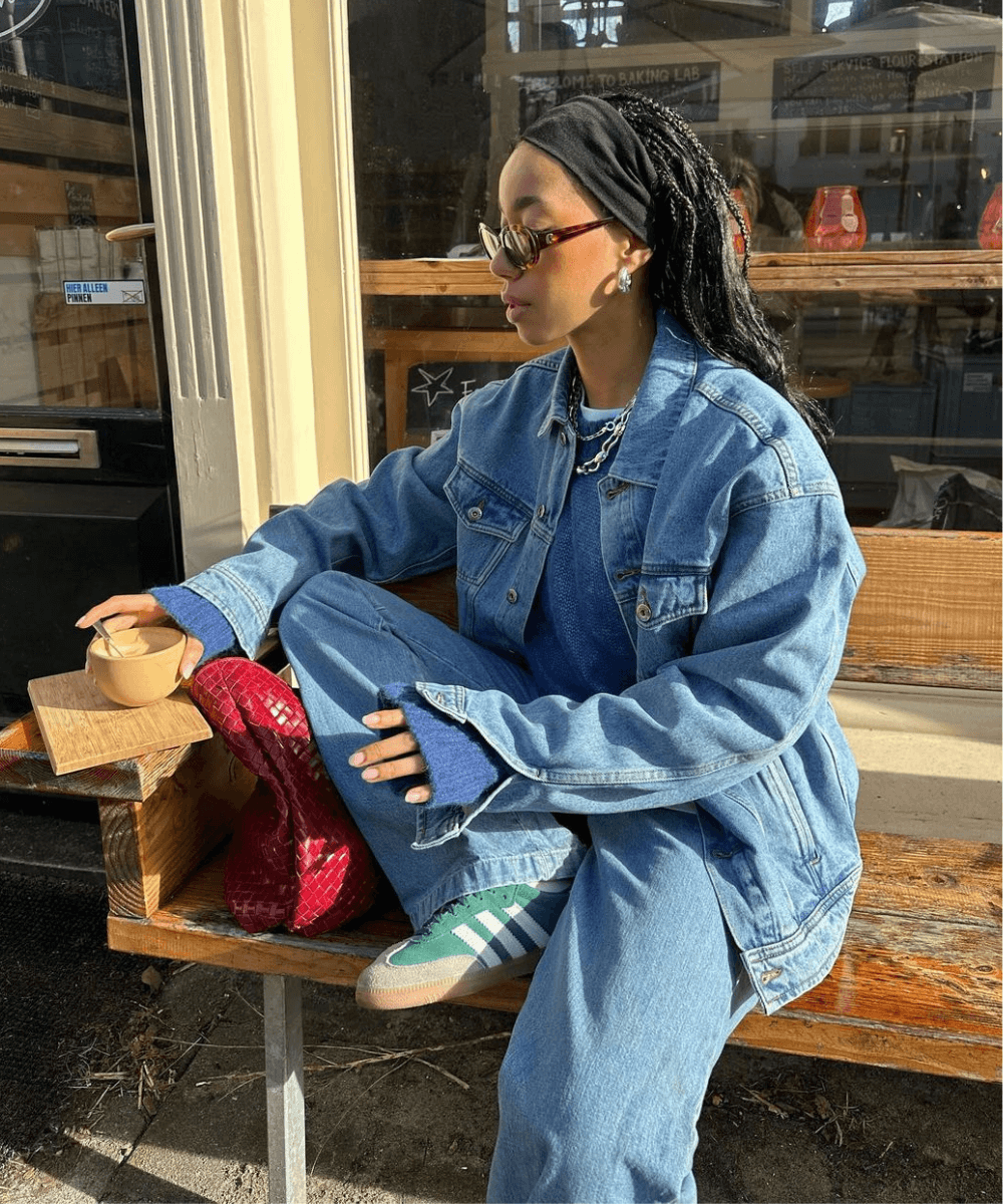 Amaka Hamelijnck - conjunto jeans e tênis adidas verde - sapatos tendência - inverno - mulher negra de óculos sentada em um banco na rua com uma das pernas dobradas - https://stealthelook.com.br