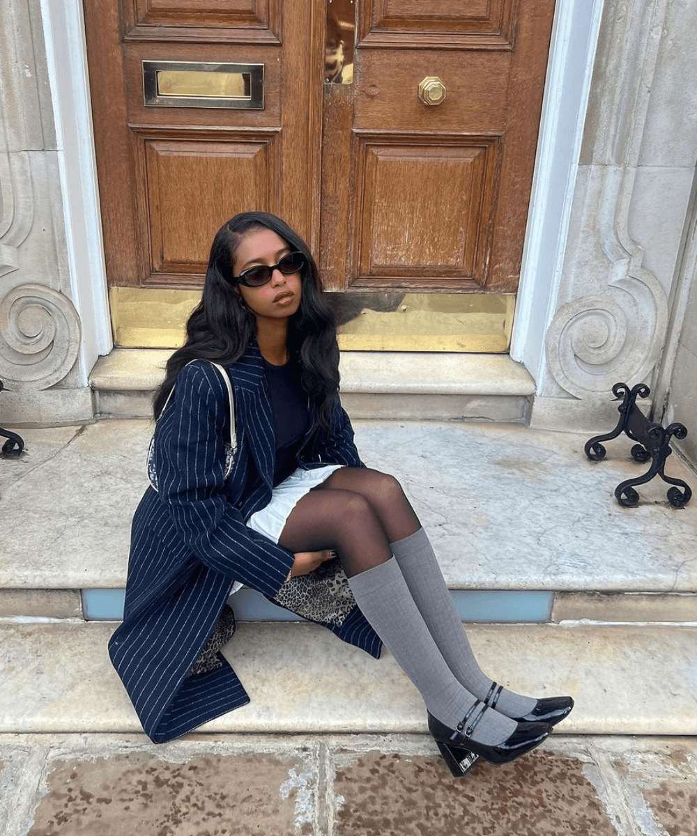 Samira Ahmed - minissaia branca, blusa preta, meia-calça e sobretudo listrado - sapatos tendência - inverno - mulher negra de óculos sentada na rua com as duas pernas dobradas - https://stealthelook.com.br