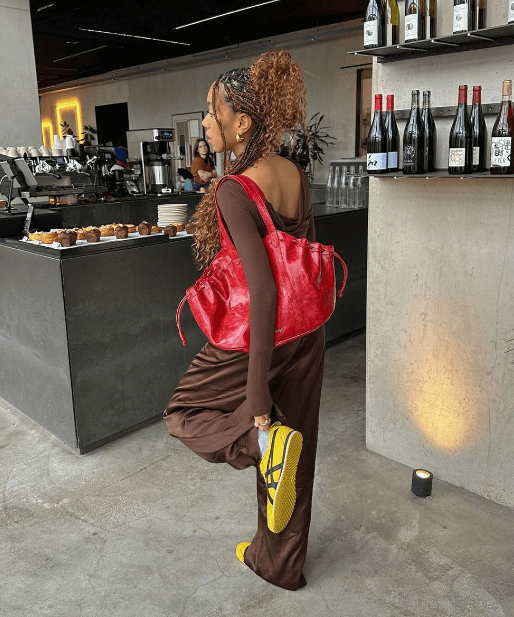 Amaka Hamelijnck - calça marrom, blusa de manga longa marrom, tênis amarelo e bolsa vermelha - cool girls - outono - mulher negra em pé de costas com uma das pernas levantada - https://stealthelook.com.br