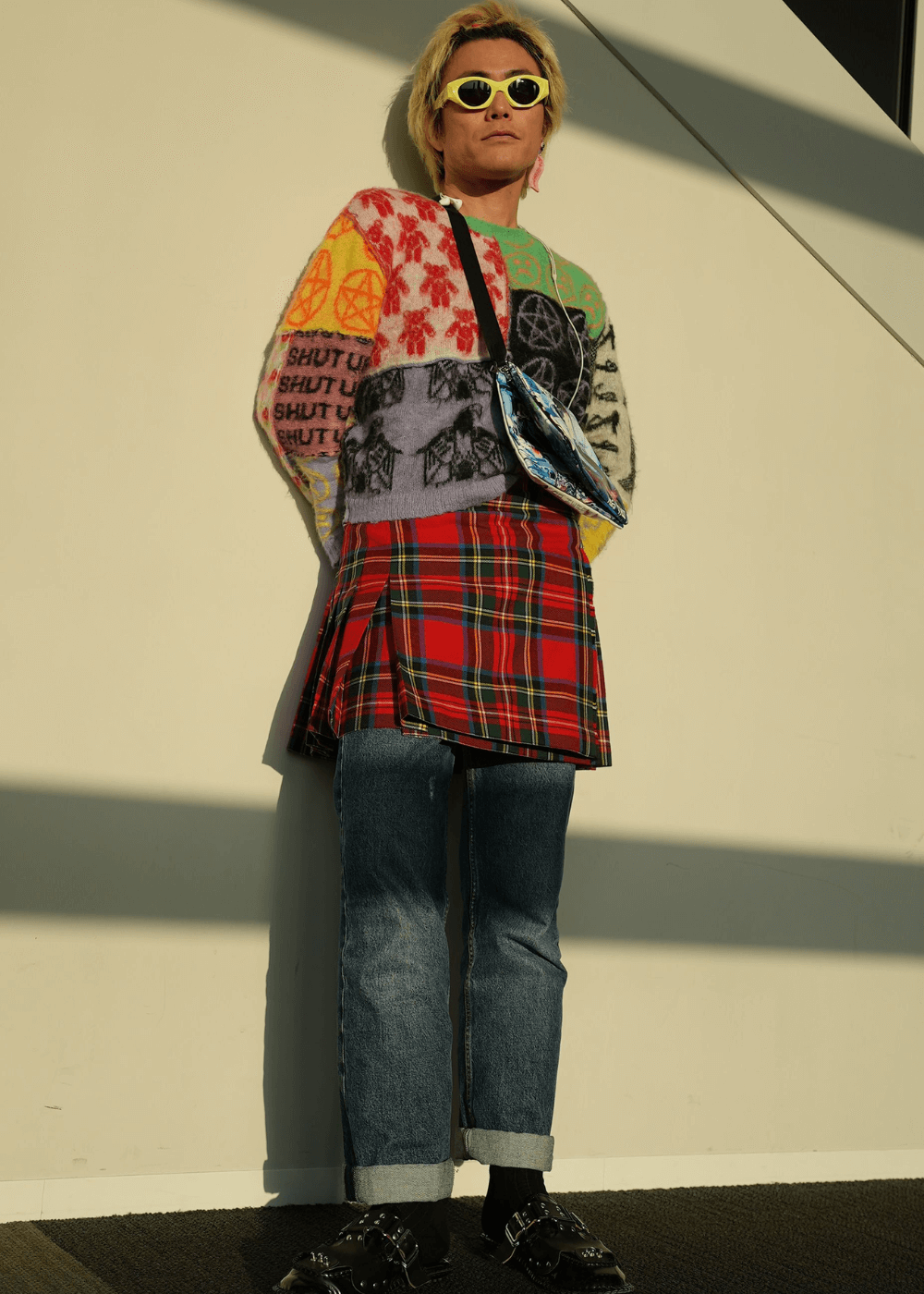 Street Style asiático - calça jeans, minissaia xadrez, tricot colorido e óculos amarelo - Street Style asiático - inverno - homem asiático loiro usando óculos de sol parado em pé encostado em uma parede - https://stealthelook.com.br