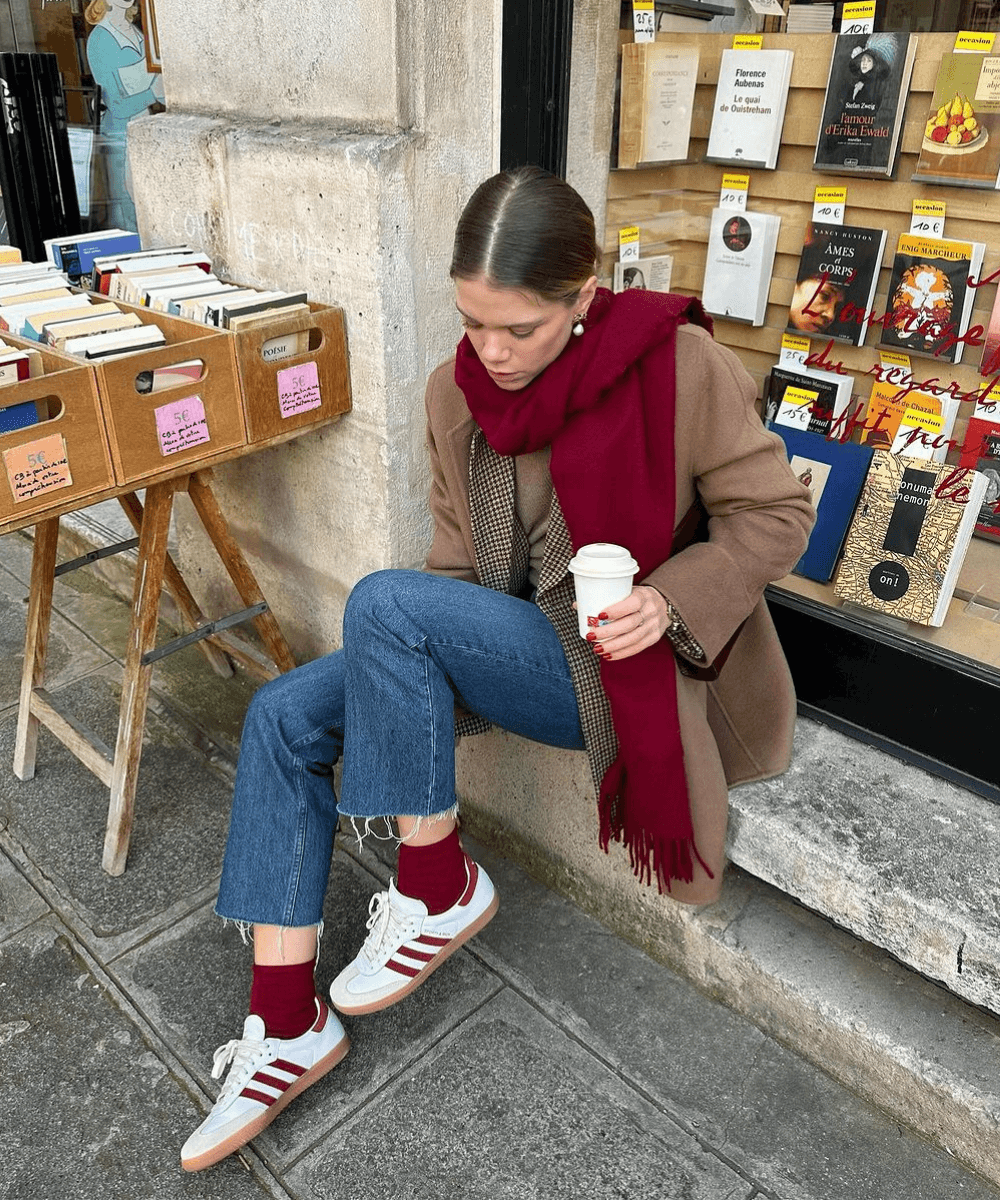 Elodie Romy - calça jeans, tênis adidas, sobretudo bege, cachechol vermelho e bolsa - como usar sobretudo - inverno - mulher loira sentada na rua - https://stealthelook.com.br