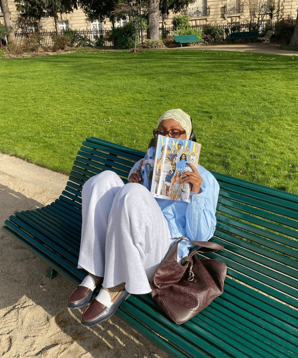 @blackmaroccan - calça, camisa azul, sapatilha e bolsa marrom - cores de bolsa - outono - mulher negra sentada em um bando no parque segurando uma revista na frente do rosto - https://stealthelook.com.br