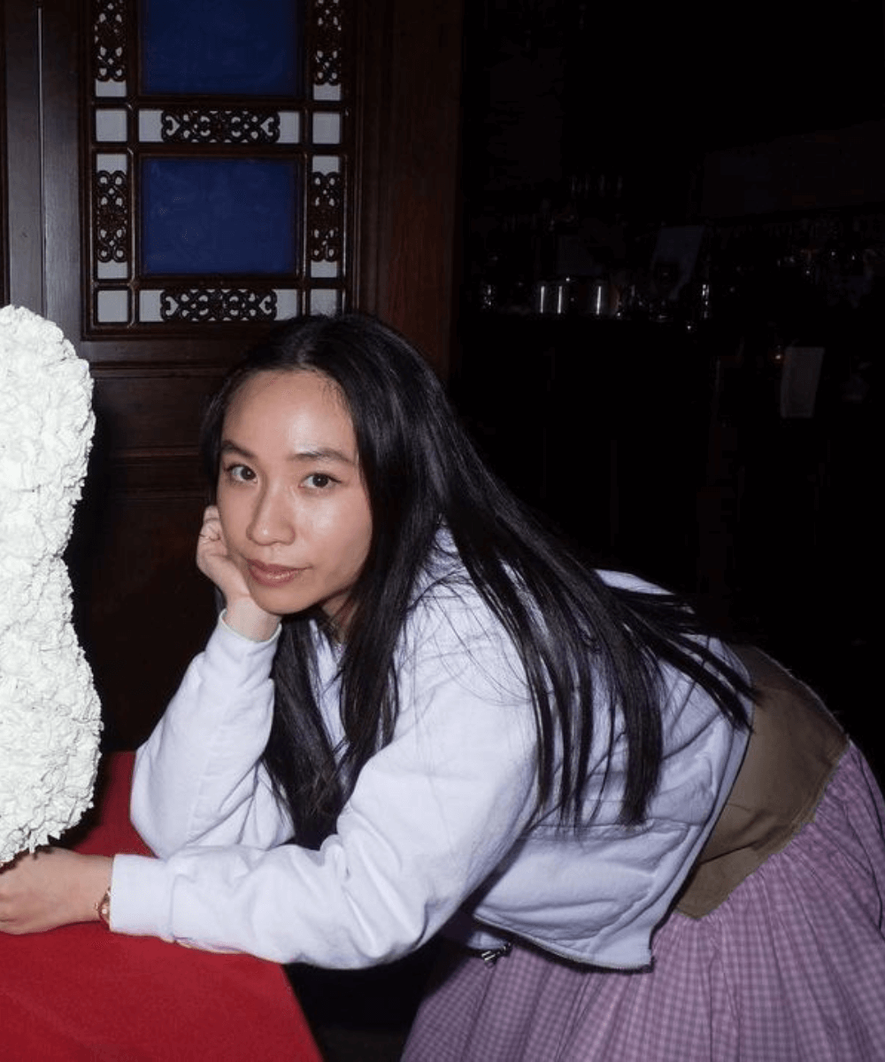 Sandy Liang - saia rosa e blusa de manga longa branca - Designers asiáticos - outono - mulher asiática debruçada sob uma mesa - https://stealthelook.com.br