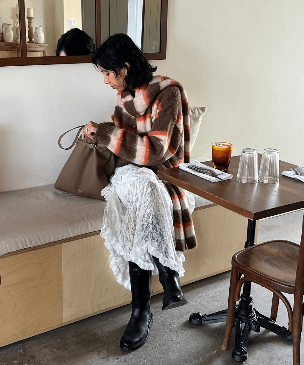 Stephanie Arant - saia midi branca, trico listrado e bota cano longo preta - looks de frio - inverno - mulher sentada em um banco dentro de uma loja - https://stealthelook.com.br