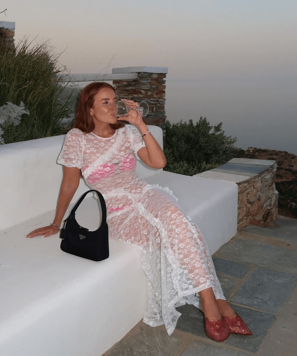 @rebeccaferrazwyatt - vestido longo branco de renda, conjunto rosa e sandália rosa - looks transparentes - verão - mulher ruiva de óculos sentada em um banco - https://stealthelook.com.br