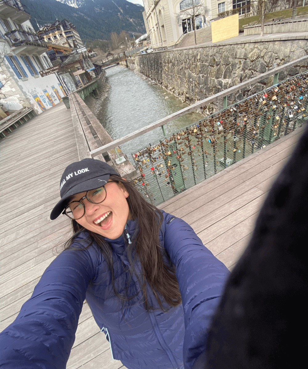 Iza Suzuki - jaqueta azul de nylon e boné preto - neve - inverno - mulher asiática tirando selfie na frente de uma ponte com cadeados - https://stealthelook.com.br