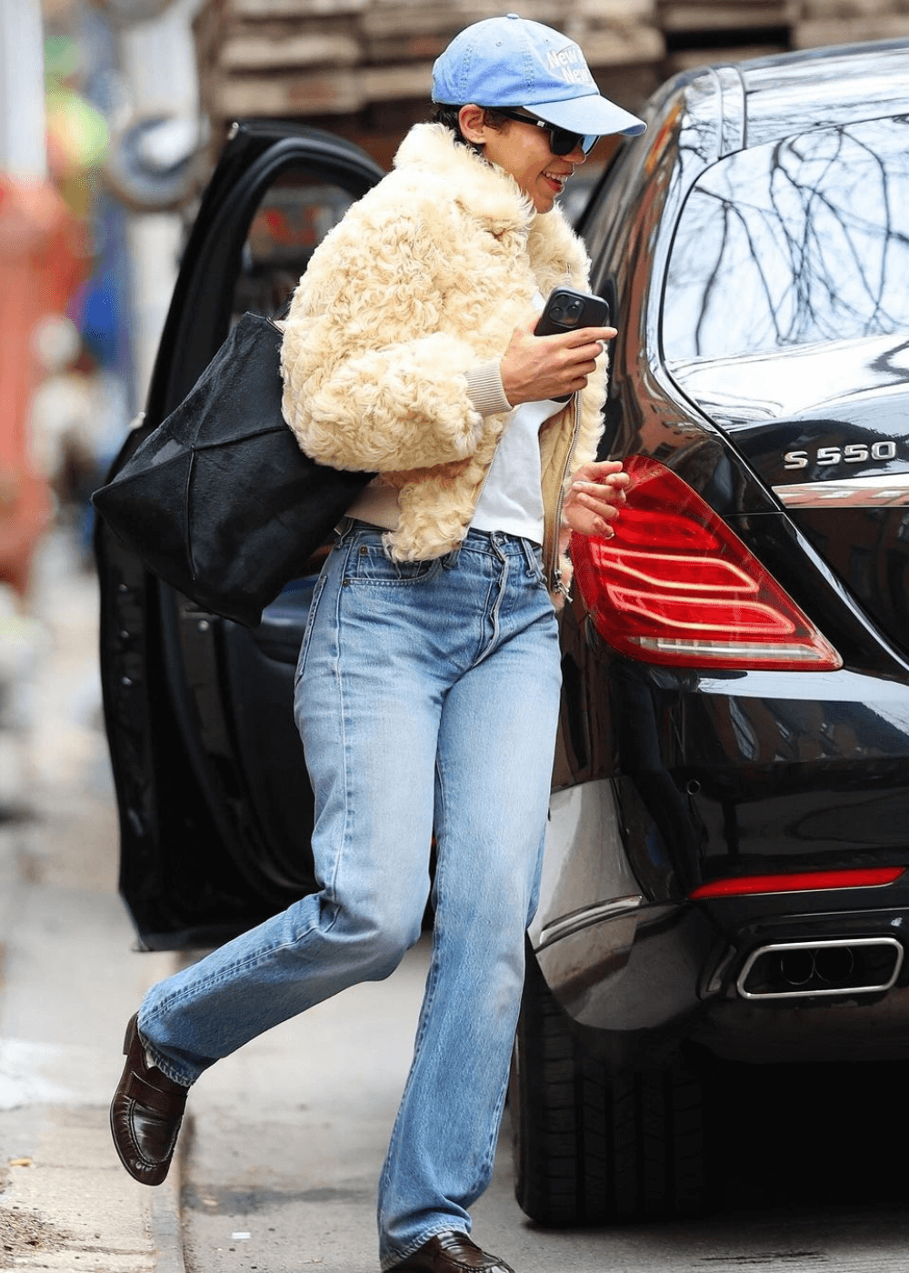 Taylor Russell - calça jeans, casaco de pelúcia, boné azul e óculos - Taylor Russell - inverno - mulher em pé na rua usando óculos de sol e um boné - https://stealthelook.com.br
