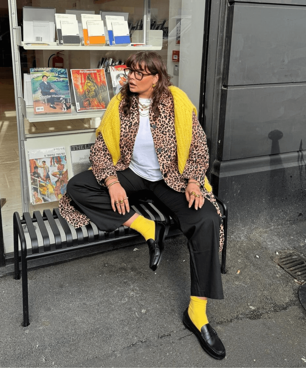 Francesca Perks - calça, meia amarela, mocassim, casaco animal print longo - outono 2024 - outono - mulher sentada em um banco na rua - https://stealthelook.com.br