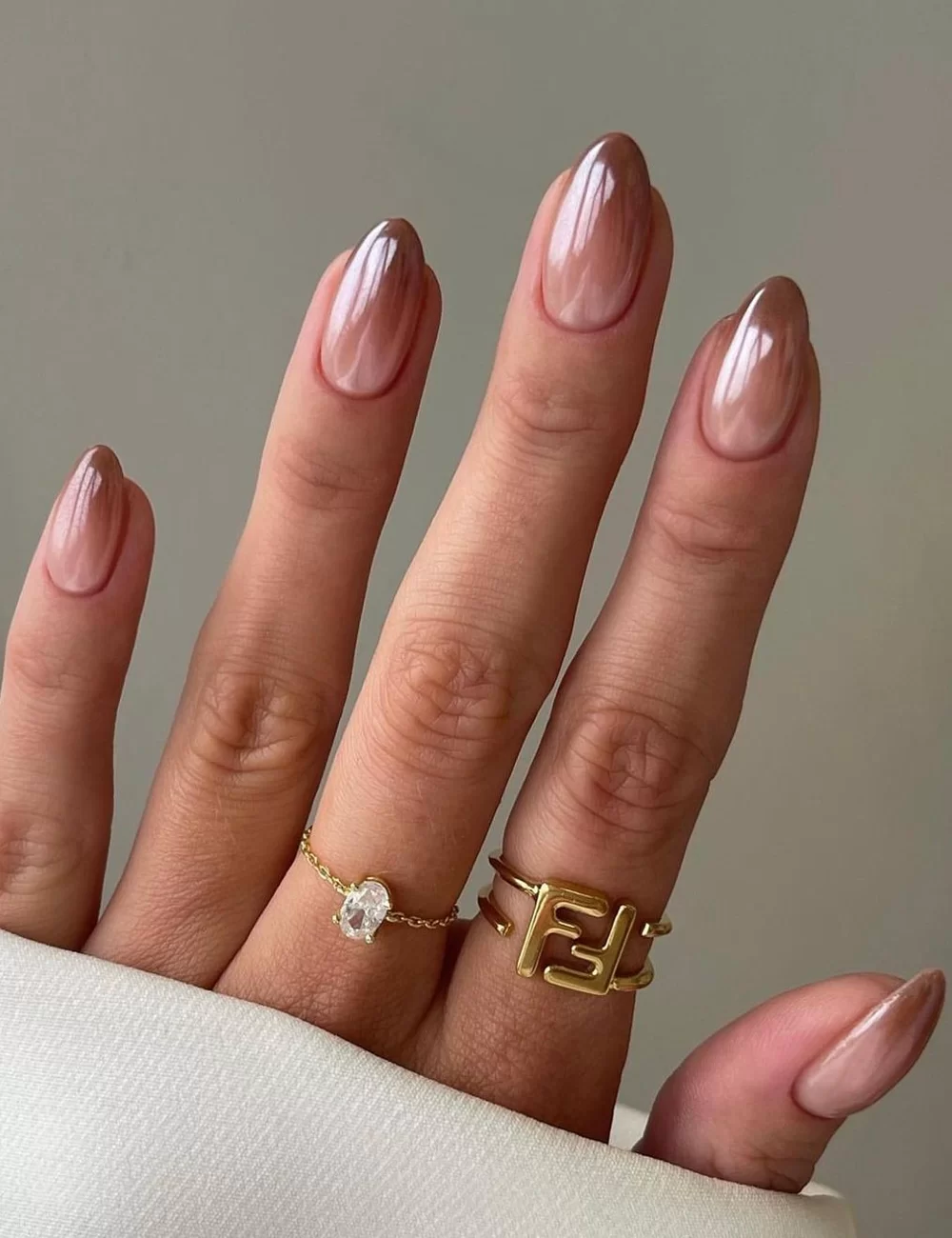 Unha amendoada com esmalte em tom nude brilhante, formato oval, combinadas com anéis delicados de ouro, incluindo um com design geométrico e outro com pedra preciosa. Elegância minimalista e tendência de moda em jóias e manicure.