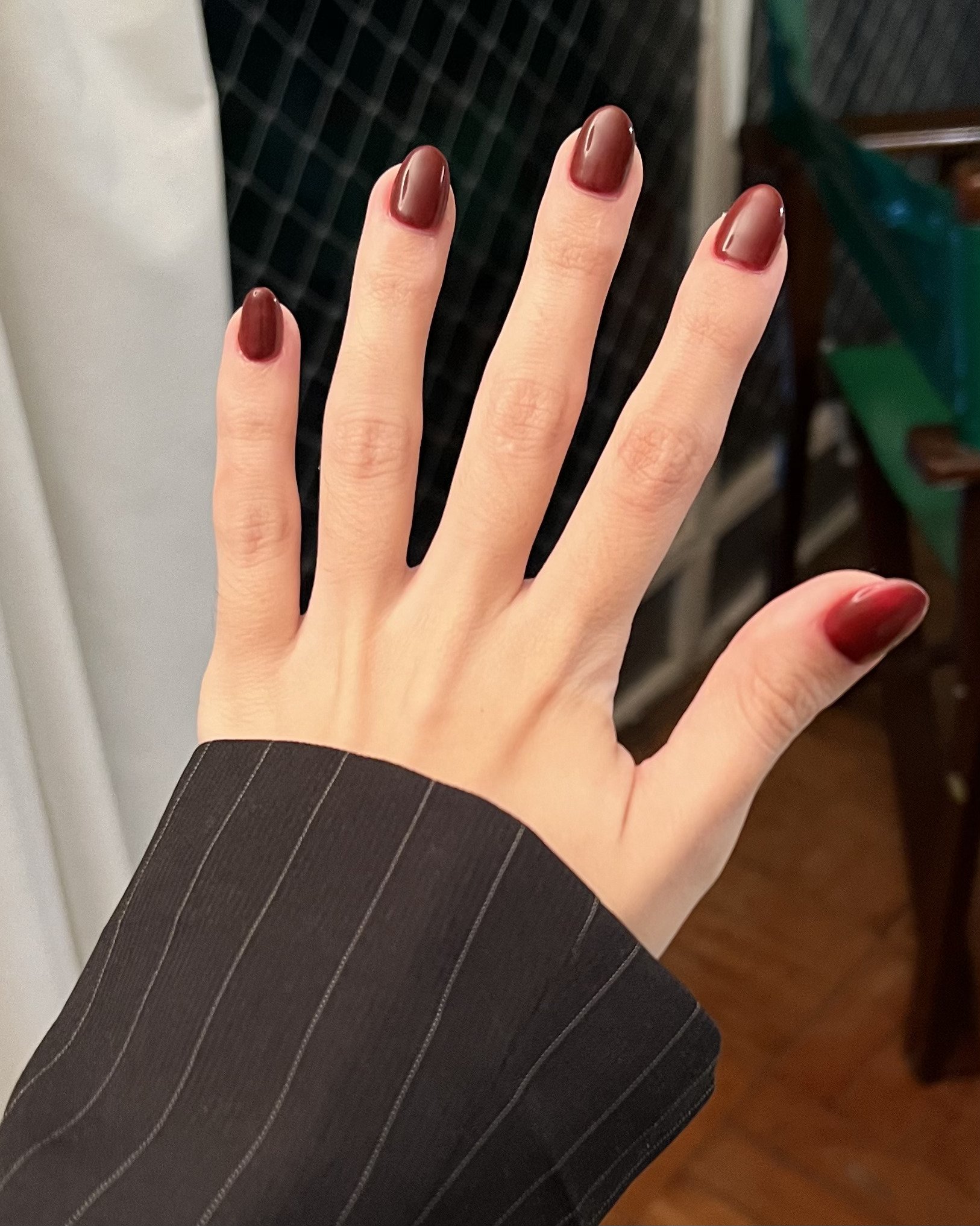 Unhas em gel amendoada pintada com esmalte vinho escuro, tendência de moda em manicure. Manga de blazer preto com riscas finas brancas, estilo formal e elegante. A combinação exala sofisticação e profissionalismo.