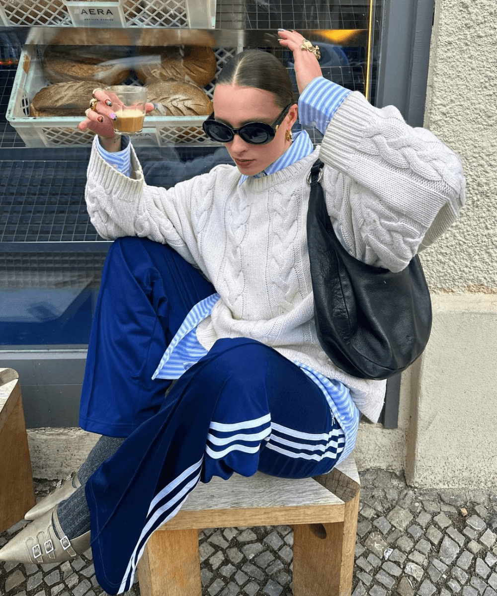 @lara_bsmnn - calça adidas azul, camis azul, tricot off white e tênis - calça esportiva - inverno - mulher sentada em um banco na rua usando óculos de sol - https://stealthelook.com.br