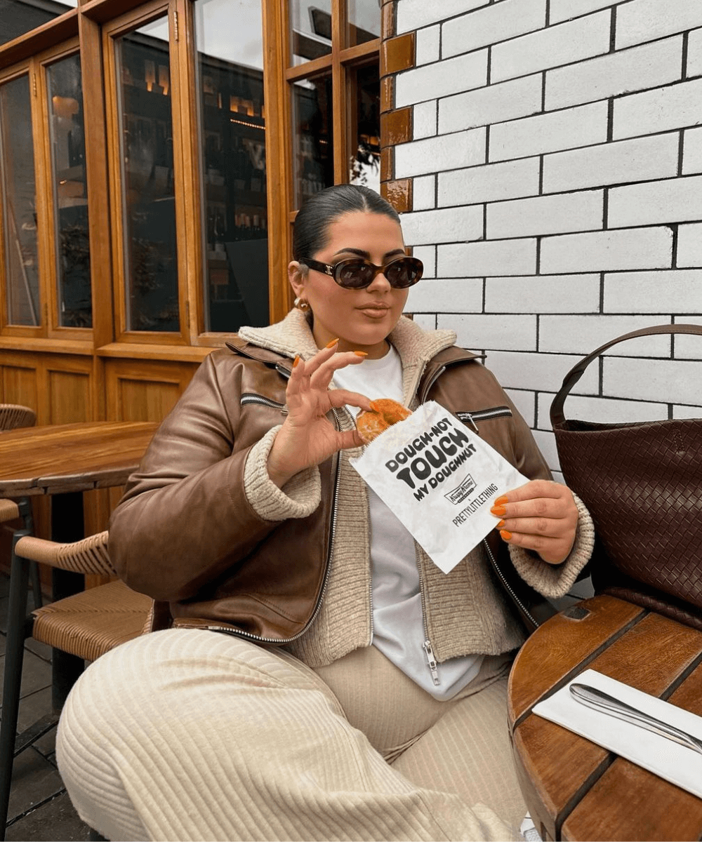 Freya Broni - conjunto de tricô, óculos oval, jaqueta de couro marrom - óculos tendência - inverno - mulher sentada em um banco na rua usando óculos de sol - https://stealthelook.com.br