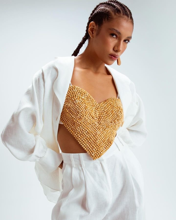 Modelo com tranças vestindo top de coração texturizado amarelo, blazer branco oversized e calça solta branca de cintura alta. Look contemporâneo que combina conforto com elementos estruturais, refletindo tendências de moda sustentável e artesanal.