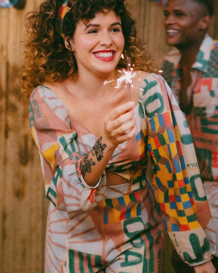 Pessoa vestindo um conjunto colorido e estampado, com influências retrô e boho, composto por blusa de mangas bufantes e calça larga. Tatuagens no braço e acessório de cabelo combinando completam o visual moderno e descontraído.