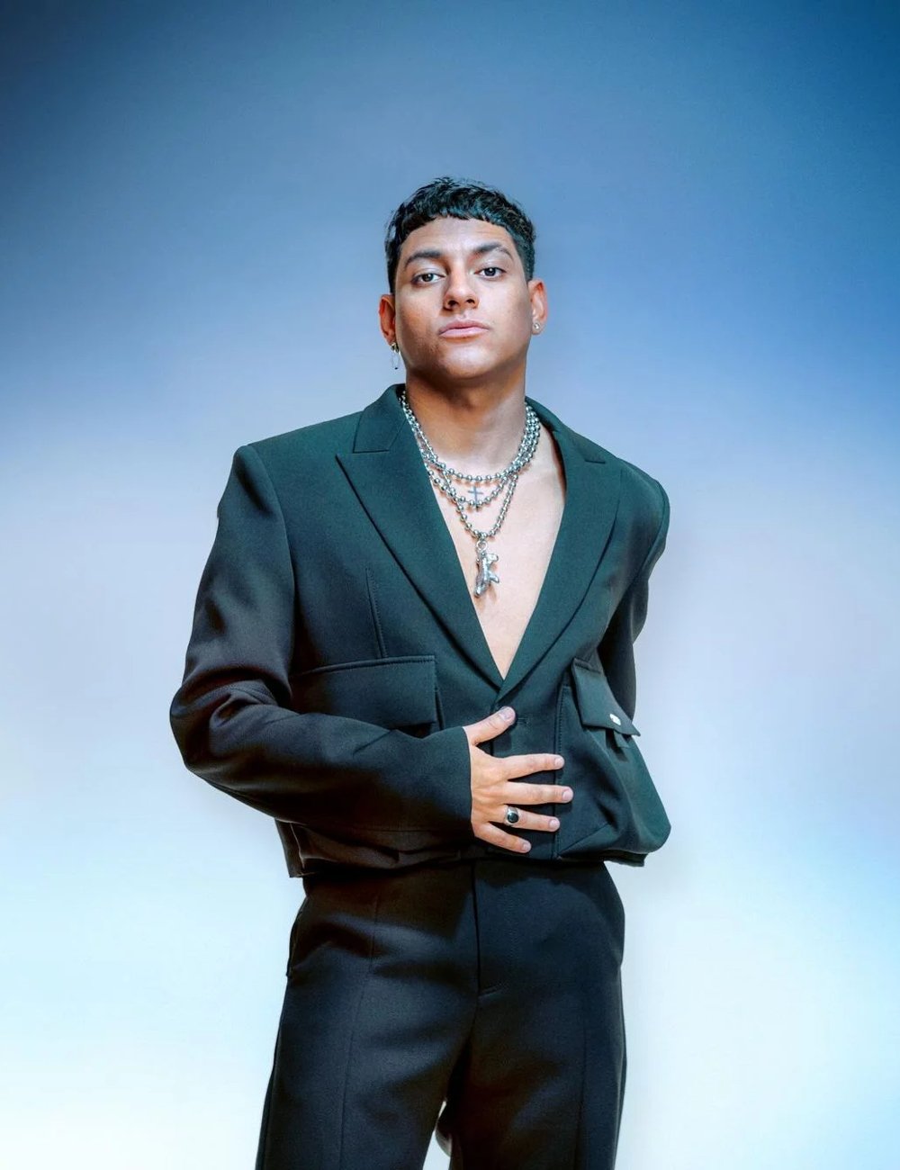 Homem vestindo um blazer preto oversized sem camisa, completando o visual com colares de corrente prateados e um anel preto. O estilo reflete tendências contemporâneas de moda, combinando elementos de elegância e ousadia. Playlist do Spotify.