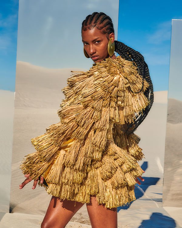 Modelo usando vestido dourado volumoso, com franjas e textura metálica, inspirado em tendências futuristas e de alta costura. Cabelo trançado com penteado elegante e brincos grandes completam o look ousado e inovador. Cenário desértico ao fundo.