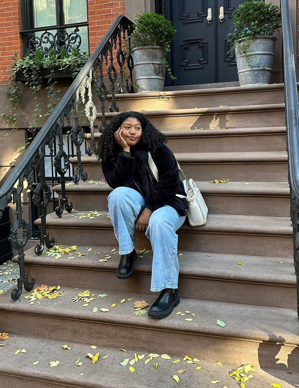 Pessoa sentada em uma escada ao ar livre usando um casaco de pelúcia preto, calça jeans larga e botas pretas de cano curto. Estilo casual e confortável, com toques de moda urbana e contemporânea das estações frias. Um look despojado e estiloso.