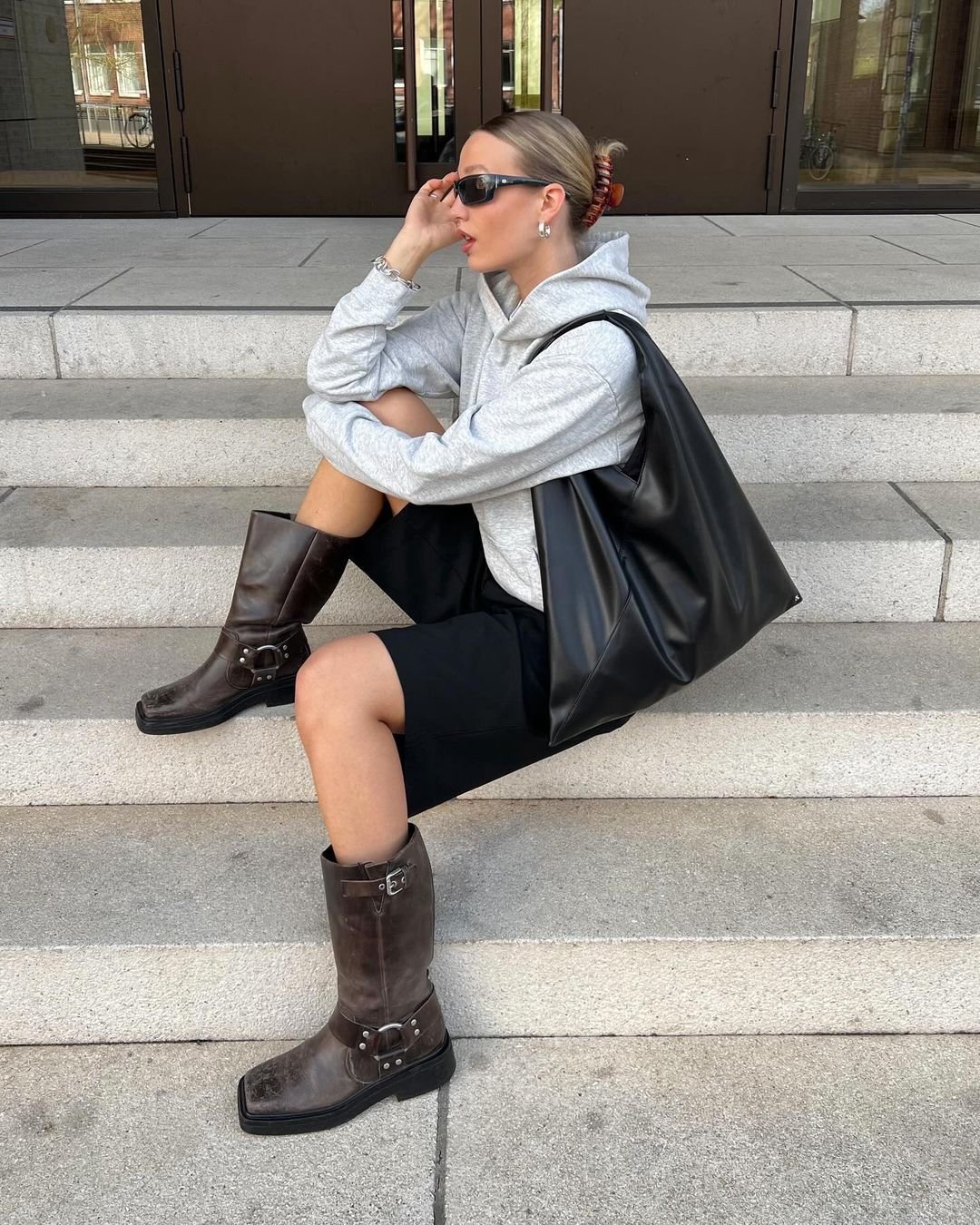 Mulher com estilo casual urbano: moletom cinza, shorts preto, bota de couro marrom, bolsa grande preta, óculos escuros e acessórios prateados. Cabelo preso com um coque e presilha. Combinação de conforto e elegância contemporânea.