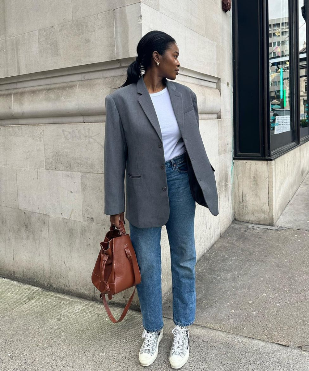 Mulher usa blazer cinza oversized, camiseta branca, jeans retos e tênis estilo casual. Completa o look com uma bolsa marrom de couro e brincos delicados. Estilo moderno e despojado, tendência streetwear com toques clássicos e confortáveis.