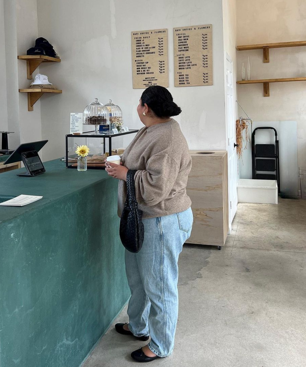 Mulher com suéter bege oversized, calça jeans de lavagem clara e sapatilhas pretas em um café minimalista. Tendências de moda incluem conforto e casualidade com peças básicas e soltas. Bolsa preta trançada completa o visual.